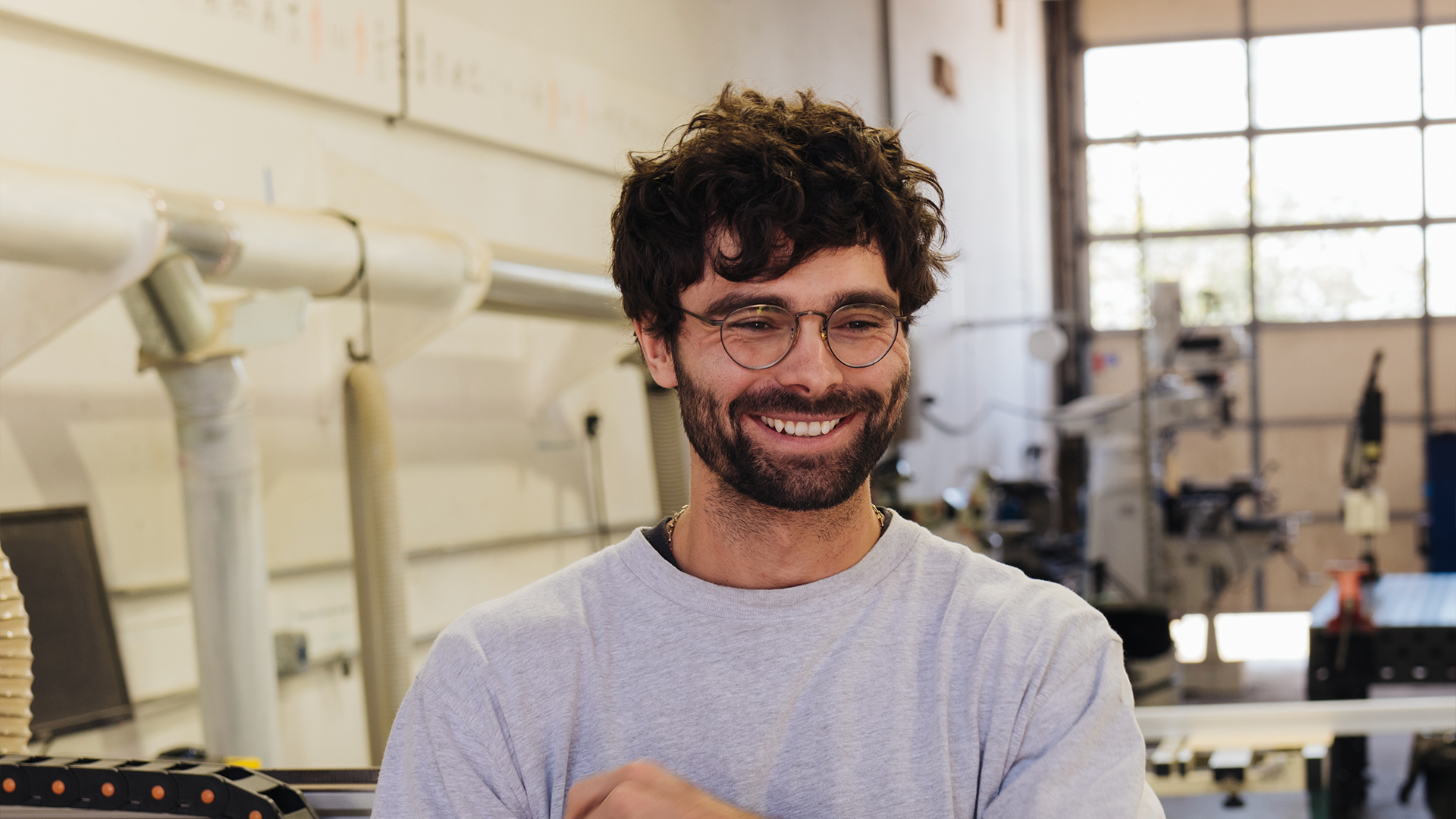 Michael Kozlowski smiling in glasses and a grey sweatshirt in a workshop full of machines
